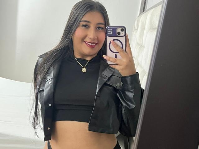 Catalina Rivero, avec ses longs cheveux noirs, souriante, portant un crop top noir et une veste en cuir, tenant un téléphone avec un support pour anneau, montrant son ventre et un collier en or..