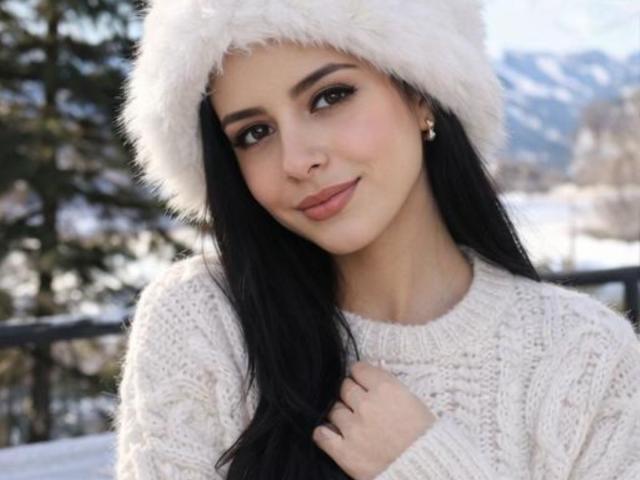IsabellaJames with long black hair, wearing a fluffy white hat and cream knit sweater, smiling softly, holding her hair. Snow-covered background, mountains, and trees in the blur.