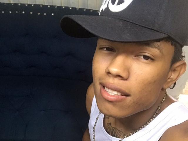 A selfie of JuanmHata with a medium-dark skin tone, short curly hair, and a black cap with a partial white design. JuanmHata wears a white tank top and a silver chain necklace. He has a subtle smile, showing slightly parted lips, and a small tattoo on his chin. The background features a dark blue, tufted headrest. The image is focused on JuanmHata's upper body and face. The cap is slightly worn, and his expression is relaxed.