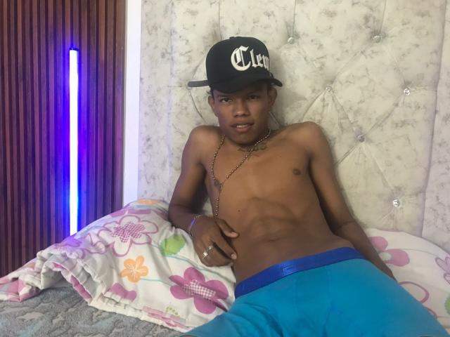 JuanmHata is a young, dark-skinned man with a lean, muscular build, sitting on a bed. He wears a black baseball cap with white text, blue boxer shorts, and a gold chain. His right hand rests on his lower abdomen. The bed has a floral-patterned pillow and a tufted, silver-gray headboard. A vertical purple LED light is on the left. His expression is neutral, and he looks slightly to the right. The image is brightly lit, highlighting his defined abs and smooth skin.