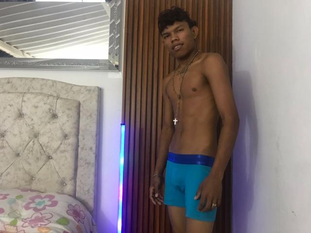 JuanmHata stands in a room, shirtless, wearing blue boxer shorts with a dark blue waistband. He has a slim, toned physique and dark brown skin. He wears a long necklace with a cross pendant. His short, curly black hair is slightly messy. He stands near a bed with a tufted, beige headboard and a floral-patterned blanket. The background features a wooden panel and a mirror. JuanmHata's expression is neutral, and he looks slightly to the right. The image is casual and unfiltered.