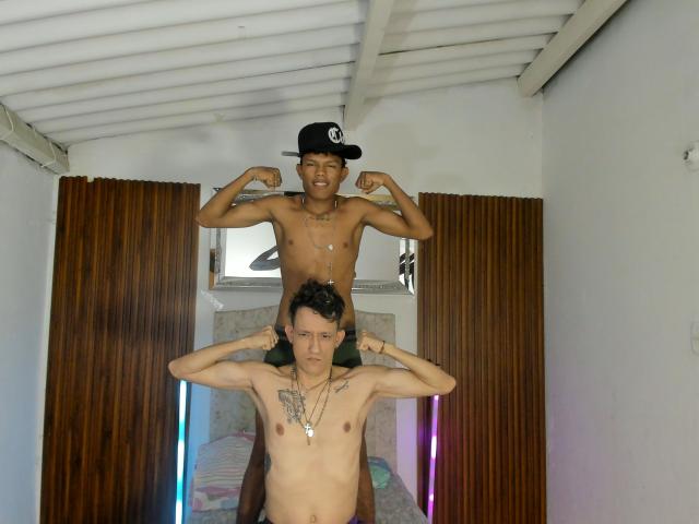 JuanAndLuke, shirtless, standing with flexed biceps, black cap, smiling, behind JuanAndLuke, also shirtless, tattoos on chest, necklace, hands on head, white ceiling, brown curtains.