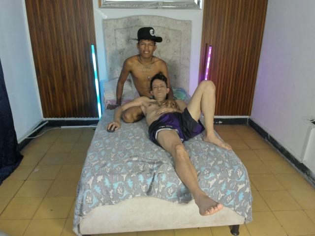 JuanAndLuke, shirtless, wearing black cap and shorts, sits behind Luke, shirtless, with purple shorts, on a bed with patterned blue and gray cover. Neon lights on walls.