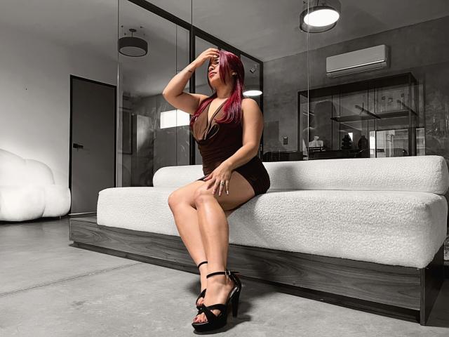 JessyRabbit with red hair, wearing a dark brown dress, black high heels, sitting on a white couch, hand on head, legs crossed, showing her ass and tits.