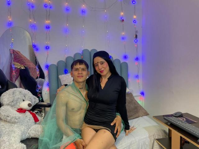 AnjaliRohan, a slim, light-skinned man with short brown hair, wears a sheer green mesh top and orange shorts. He stands beside AnjaliRohan, a medium-skinned woman with long black hair, in a black, form-fitting dress. They are on a bed with a blue, padded headboard. A fluffy white teddy bear with a red ribbon is on the left. Blue and purple fairy lights hang in the background. The woman's right hand rests on the man's hip. The image has a casual, homey feel.
