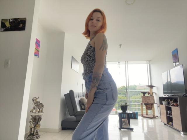SarahRousen with red hair, wearing a leopard-print top and high-waisted blue jeans, showing her tattooed arm and standing with her ass slightly raised.