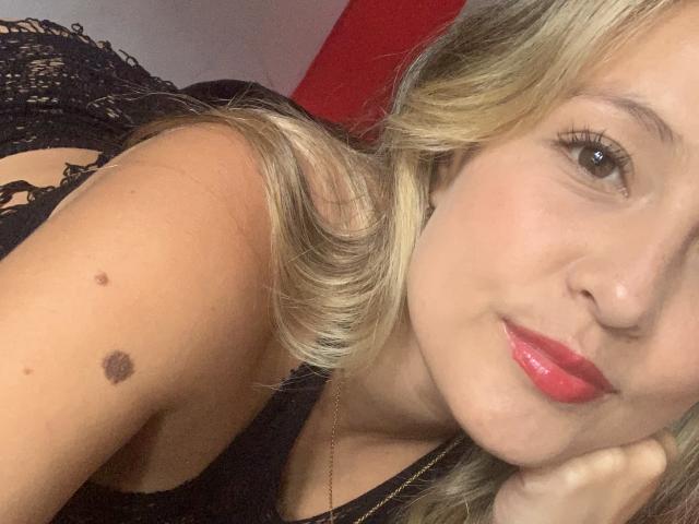 SaritaCaramelo has blonde hair, red lipstick, and a black lace top, showing a mole on her shoulder, lying with her face close to the camera.