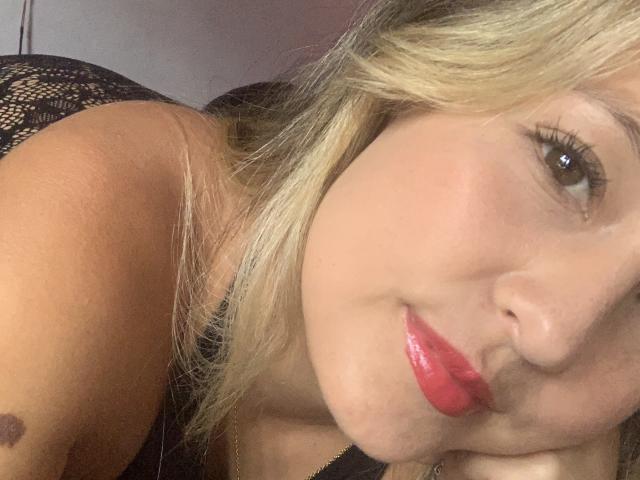 SaritaCaramelo's face, blonde hair, red lipstick, and light skin, lying down with a black lace top, small dark mole on her right cheek, soft smile, brown eyes.