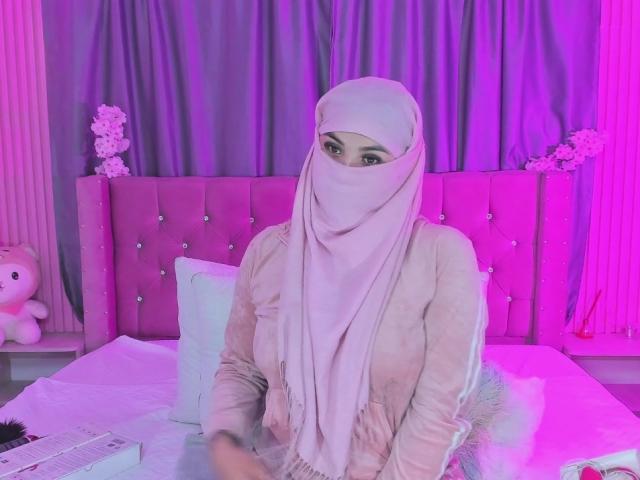 MalakCote with pale pink hijab, matching outfit, sitting on a bed with pink headboard, looking at camera, purple and pink lighting, white pillow, stuffed teddy on left.