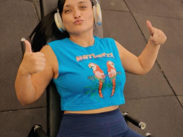 DanyQueenX, wearing a blue sleeveless crop top with two colorful parrots on the front, is seated on a gym machine. She has puffy lips and is giving a thumbs-up with her right hand, while wearing white headphones. Her dark hair is pulled back, and she's wearing dark blue pants. The texture of the crop top is smooth, and the parrots have bright, detailed feathers. Her skin is light, and she looks relaxed. The machine has black handles and a padded seat.