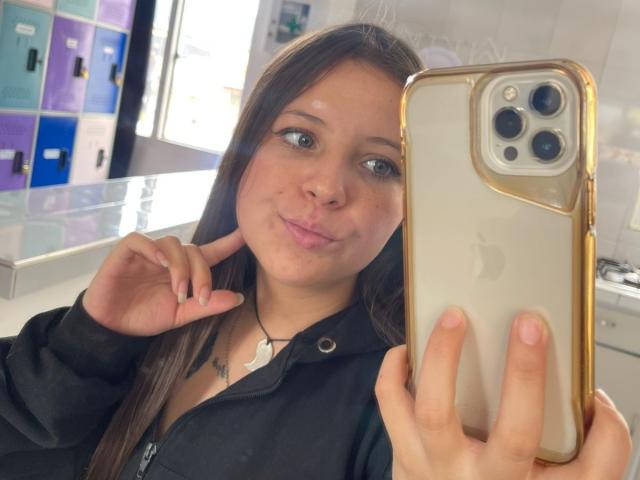GraceRedMiller takes a selfie with a gold iPhone in a bright room. She has long brown hair, light skin, and is making a playful pout with her lips. She wears a black jacket with a white shirt peeking out. Her right hand holds the phone, and her left hand points to her cheek. The phone case is gold with a white back. The background includes colorful locker panels. The image is clear with natural light.