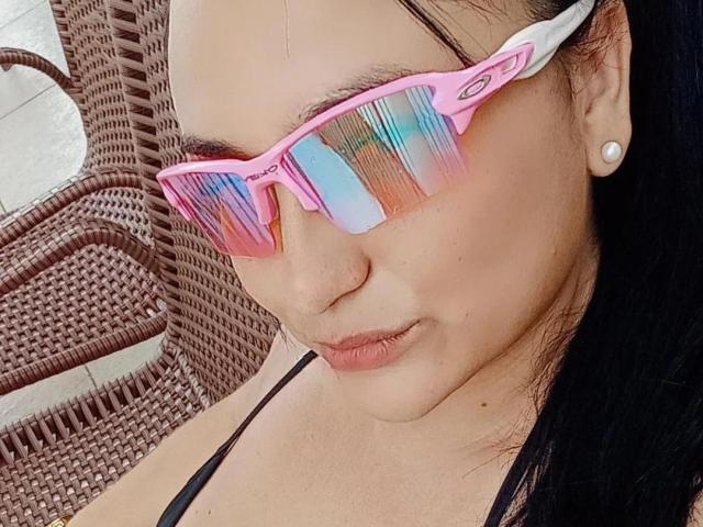 DanyQueenX usando óculos de sol com efeito degradê rosa, cabelo preto preso em um rabo de cavalo, e um brinco de pérola, exibindo um nariz delicado e um sorriso discreto, vestindo uma blusa preta com alças..