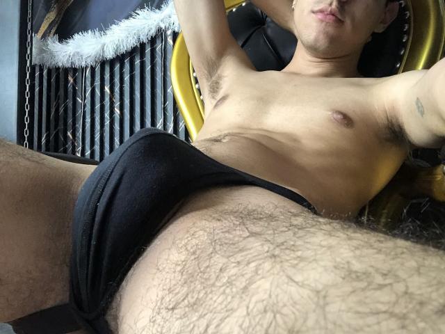 NicholasFinn, shirtless, wearing black underwear, lying back with arms raised, showing a hairy chest and a hairy, slightly exposed, crotch. His face is partially visible, with a pouty expression.