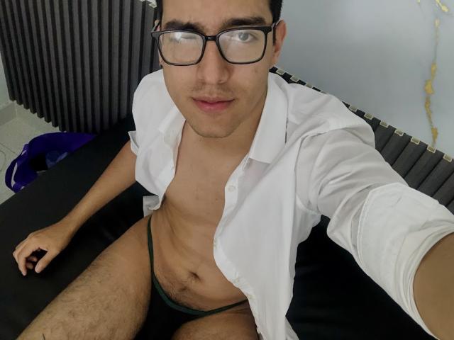 NicholasFinn with glasses, white shirt open, showing a slim, hairy chest, and green jockstrap, sitting on a black bed, holding a selfie stick.