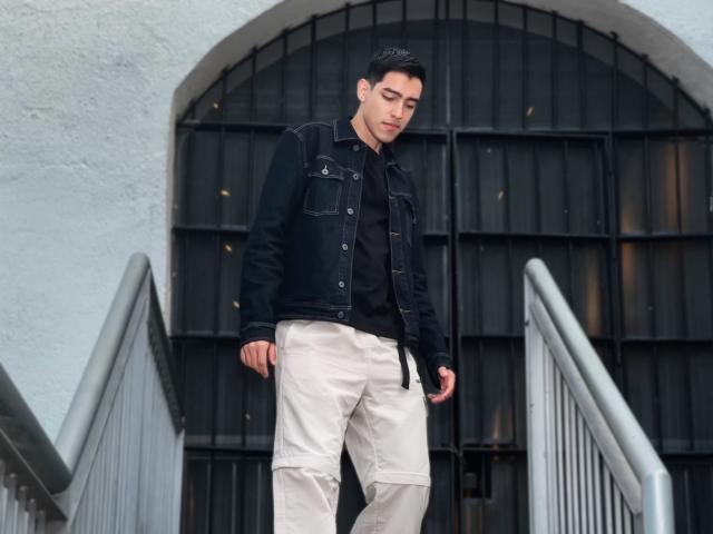 NicholasFinn, wearing a black denim jacket over a black shirt, and beige pants, standing on a staircase with a black arched door in the background.