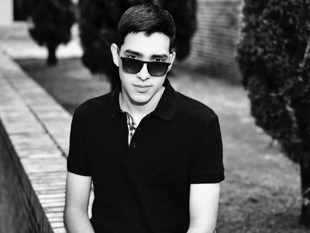 Black and white photo of NicholasFinn with short dark hair, wearing sunglasses and a black polo shirt, standing with a slight smile, showing a cute ass.
