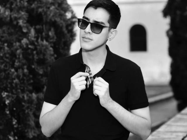 NicholasFinn with dark sunglasses, short black hair, black polo shirt, holding a plaid tie, looking slightly to the side, standing in front of a blurry building.