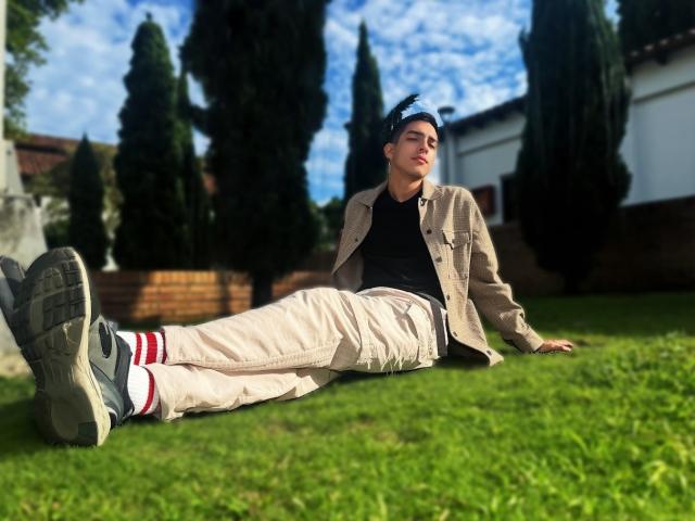 NicholasFinn is lying on grass, wearing a beige jacket, black shirt, white pants, grey sneakers with red stripes, and a black cap, with his ass in the air.