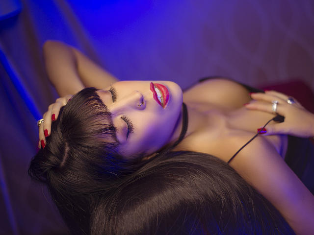 SeleneBright with her eyes closed, head tilted back, long black hair, red lipstick, and a black choker. Her hands are on her head, showing red nails and a ring.