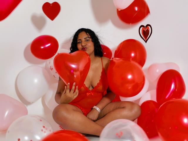 LiliLopez, with dark curly hair and red lingerie, holds a red heart balloon, surrounded by red and white balloons, sitting cross-legged, displaying her tits and cute face.