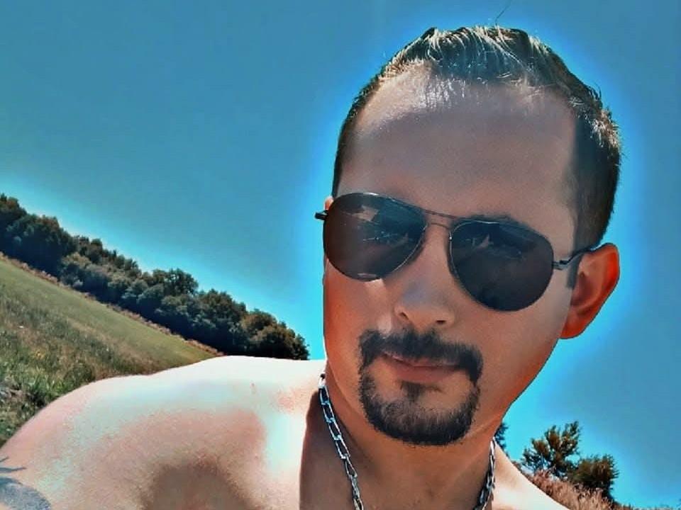 Close-up of KikoTop, a shirtless man with a short, dark beard and mustache, wearing aviator sunglasses and a chain necklace. His hair is slicked back, and he has a tattoo on his left shoulder. The bright blue sky and green, grassy hill with trees in the background are blurred. KikoTop's skin is tan, and he has a confident, slightly smug expression. The image has a glowing, halo-like effect around his head.