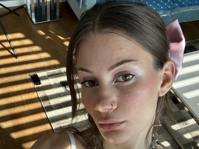 CassyCox has brown eyes, pink nose ring, light pink lipstick, pink bow, and white tank top, with shadowed wood floor and striped rug in a sunlit room.