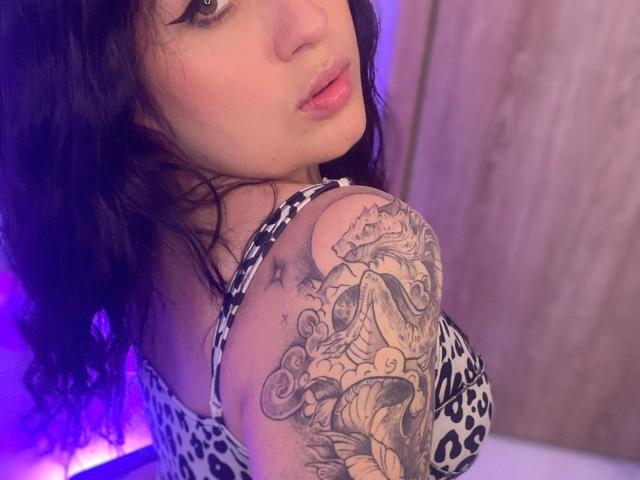 AnnyBentleii, con cabello negro ondulado, mirando hacia atrás, mostrando un tatuaje detallado de un dragón y olas en su hombro derecho, vestida con un vestido de estampado de leopardo, labios rosados..