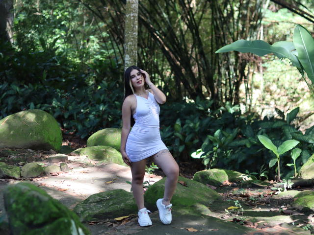 KatieDesirX stands in a forest, wearing a white, ruched, sleeveless dress and white sneakers, with her hand in her brown hair, showing her ass and small tits.
