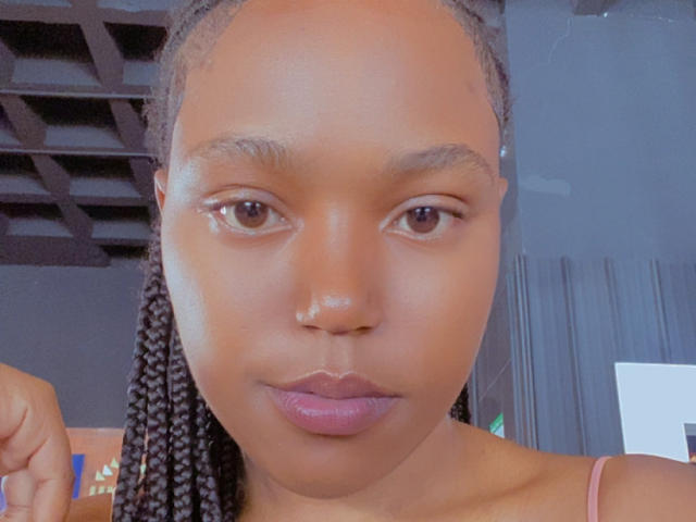 Close-up of MochiSexy, a dark-skinned woman with straight, medium-length braids. She has a small nose ring in her left nostril and wears pinkish-purple lipstick. Her brown eyes are centered, and her expression is neutral. The lighting is even, highlighting her smooth skin. She's wearing a pink strap over her shoulder. The background is blurry, with a dark ceiling and a white rectangular object on the right. The image has a slightly grainy texture.