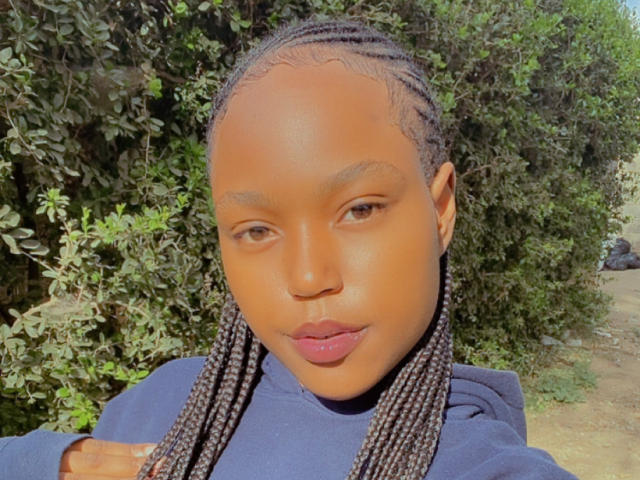 MochiSexy is a young Black woman with smooth, dark brown skin and straight, thick braids that hang over her shoulders. She wears a navy blue hoodie and has full, pink lips. Her brown eyes are slightly downcast. The light highlights her shiny skin. She's standing in front of a dense green bush, with a dirt path and a small rock visible in the background. The image is clear and detailed.