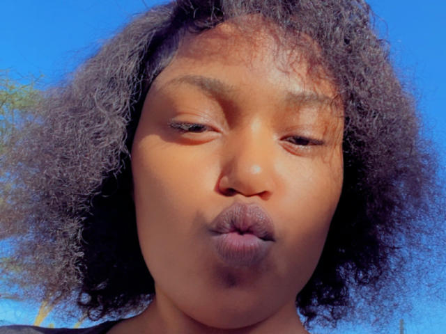 MochiSexy with dark brown curly hair, puffy lips making a kissing face, eyes half-closed, under bright blue sky, skin tone is medium brown, looking cute and playful.