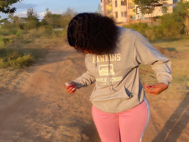 MochiSexy with curly black hair, gray hoodie, and pink leggings, standing on a dirt path, looking down at her hands, in a sunlit outdoor setting.