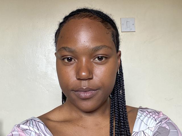 MochiSexy has dark brown skin and is looking directly at the camera with a neutral expression. They have small, slightly upturned lips and dark brown eyes. MochiSexy's hair is in tight, medium-length braids that fall straight down past their shoulders. They're wearing a white and purple striped top with puffy sleeves. A small white electrical switch is on the beige wall behind them. The image is a straightforward, realistic portrait. The texture of MochiSexy's skin is smooth with a slight sheen.