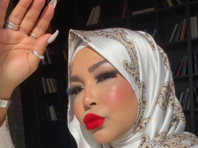 AngelinaDeleon with red lipstick, white patterned hijab, and long white nails, waving with silver rings, has a cute, chubby face, and is looking off to the side.