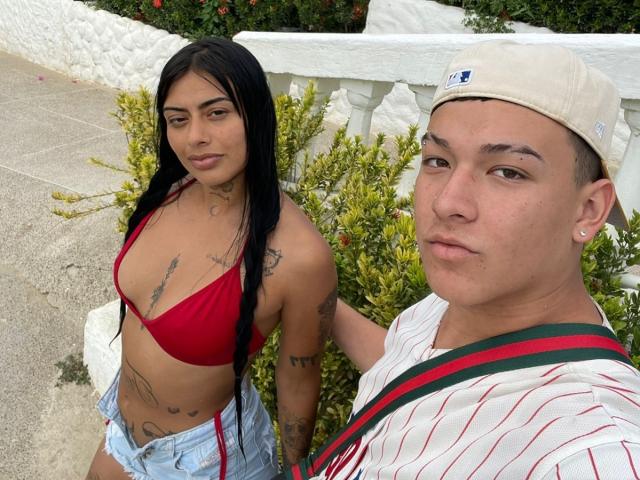 Two people stand close together in a photograph. ZairaAndres, a Latina woman with long black hair, wears a red bikini top and light blue denim shorts, revealing multiple tattoos on her arms and torso. She has a medium build with tan skin and a slight smile. To her right, a man with a shaved head, light skin, and a serious expression wears a white cap, a striped shirt with red and green stripes, and a necklace. He holds a strap over his shoulder. The man's face is closer to the camera, while ZairaAndres is slightly behind him. The composition focuses on their upper bodies.