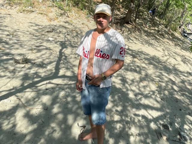 ZairaAndres stands on a sandy beach, wearing a white pinstripe baseball shirt with "Lilies" in red letters and the number 25. He's also in blue, slightly dirty, knee-length shorts and a beige cap. He's barefoot, with a gold wristwatch on his left wrist. Shadows from trees create a pattern on the sand. He's smiling slightly. Sunlight highlights his casual, relaxed posture. The image is natural and unfiltered.