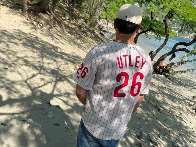 ZairaAndres stands on a sandy beach, back to the camera. He wears a white baseball shirt with red pinstripes and the name "UTLEY" along with the number "26" in red on the back. He also wears a beige baseball cap and blue jeans. His arms are behind him, and he faces a wooded area with dappled sunlight. The beach, water, and trees create a natural, serene setting.