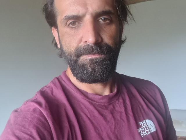 SalvaRany has a medium-length, dark, slightly wavy hair and a thick, full beard. His skin is medium brown with a few visible wrinkles around his eyes. He wears a maroon The North Face t-shirt with a small white logo on the left chest. His expression is neutral, looking directly at the camera. The light is natural, coming from the left, casting soft shadows on his face. The overall composition is centered, focusing on his upper body and face.