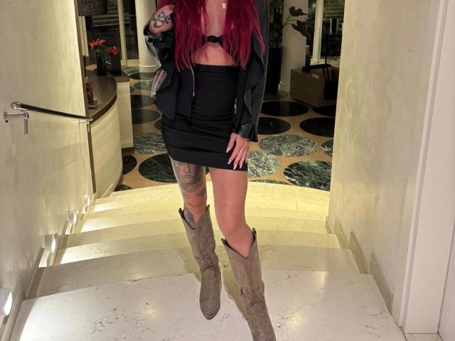 ScarletSins-hot with red hair, black strapless mini-dress, black jacket, tattoos on left thigh and arm, and tan cowboy boots, standing in a marble hallway.