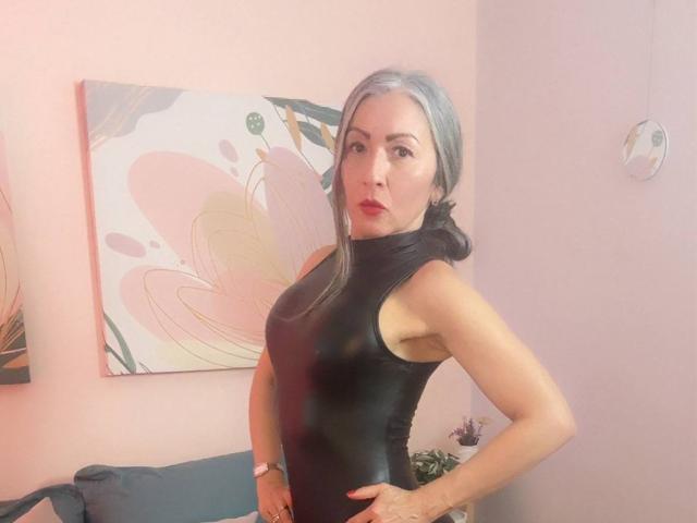 NicoletteShea with gray hair in a black latex dress, pouty lips, hands on hips, wearing a pink watch, in a pink-walled room with floral art.