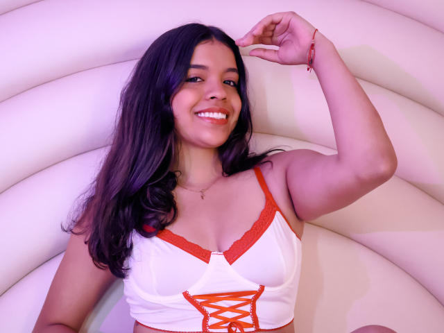 LiinaSantacruz with dark wavy hair, smiling, wearing a white lace-up top with red trim, exposing her tits, raising her right arm, and wearing a red bracelet.