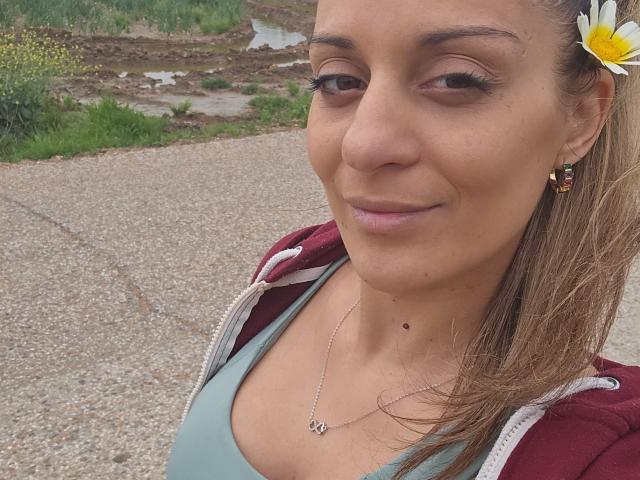 Natashha69 with light brown hair, wearing a daisy in her hair, red jacket, green top, necklace, small mole on her chin, smiling slightly, gravel road in background.