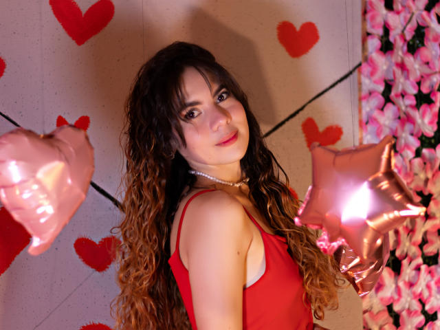 LindaPerez with curly brown hair, red dress, pearl necklace, holding pink heart and star balloons, smiling, pink flower wall, soft pink and red lighting.