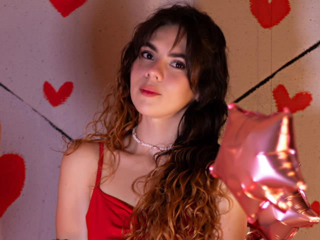 Linda Perez, avec ses cheveux bruns ondulés, une robe rouge, un collier de perles, tenant un ballon rose en forme d'étoile, regardant l'appareil photo avec un sourire discret. Ses seins sont visibles sous la robe, et elle porte du rouge à lèvres rose..