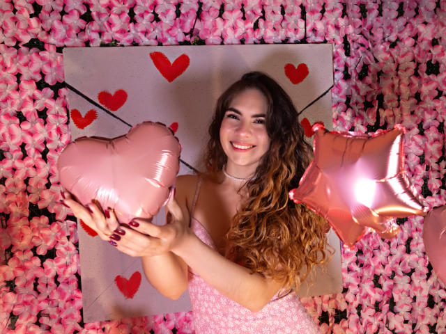 LindaPerez with curly brown hair, smiling, holding pink heart and star balloons, wearing pink dress, in front of pink flowers, has cute face, small tits, and red nails.
