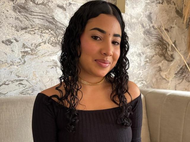 AntonelaRios with curly black hair, wearing a black off-shoulder top and gold choker, has a cute nose and a subtle smile, looking directly at the camera.