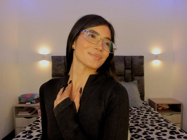 PaulethJoens with medium brown skin and straight black hair, wearing glasses with clear frames and a black ribbed shirt, stands in a bedroom. She has long black nails and a slight smile. She's centered in the frame, with her right hand near her collarbone. The bed behind her has a leopard print blanket, two bedside tables, and wall-mounted lamps. The lighting is warm and even. The image has a casual, modern feel.