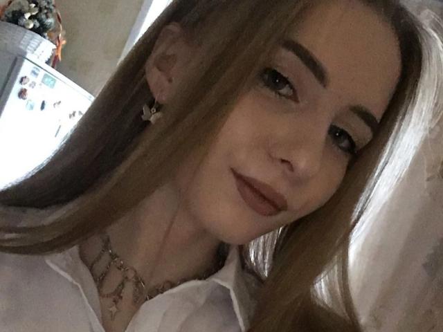 CutiCrystal with long blonde hair, dark eyebrows, and subtle makeup, wearing a white shirt and necklace, smiling softly, showing her gentle face and small nose.