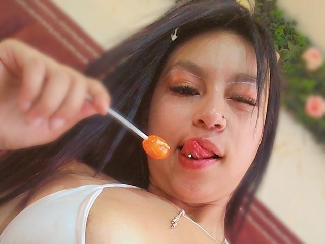 ValerieMyers with black hair, tongue out, licking an orange lollipop, has a nose piercing, and is wearing a white top, showing her upper chest and shoulder.