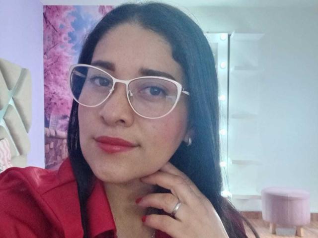 AlanaSteel with white glasses, red lipstick, and a red jacket, showing off her tits with a hand touching her cheek. She's got long black hair and a ring.